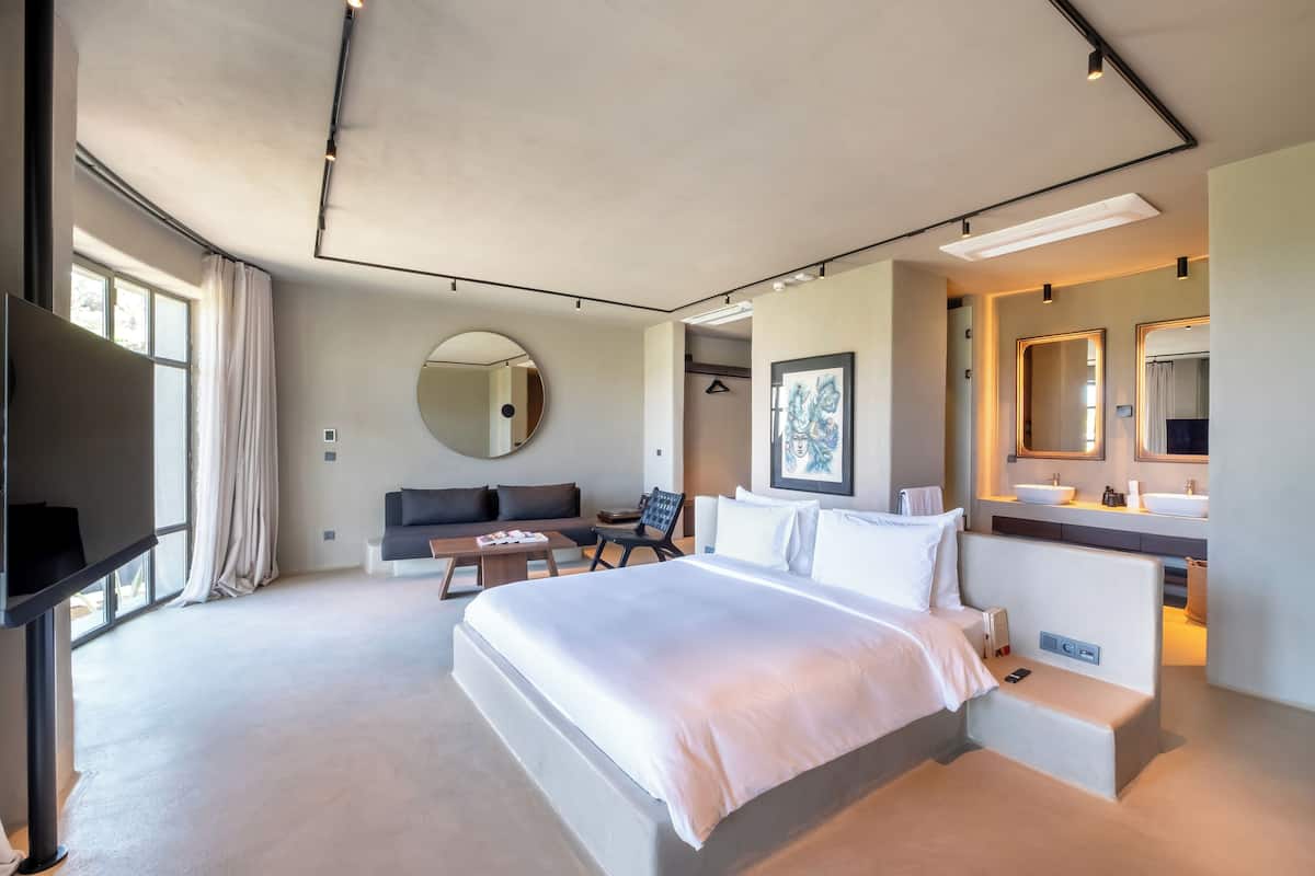 A modern hotel room with a large bed, a seating area with a sofa and chairs, a TV, and a bathroom with a mirror and sink.