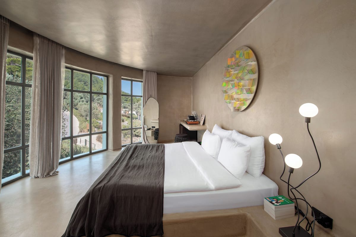 A modern bedroom with a large bed, a bedside table, a wall-mounted artwork, and a view through large windows.