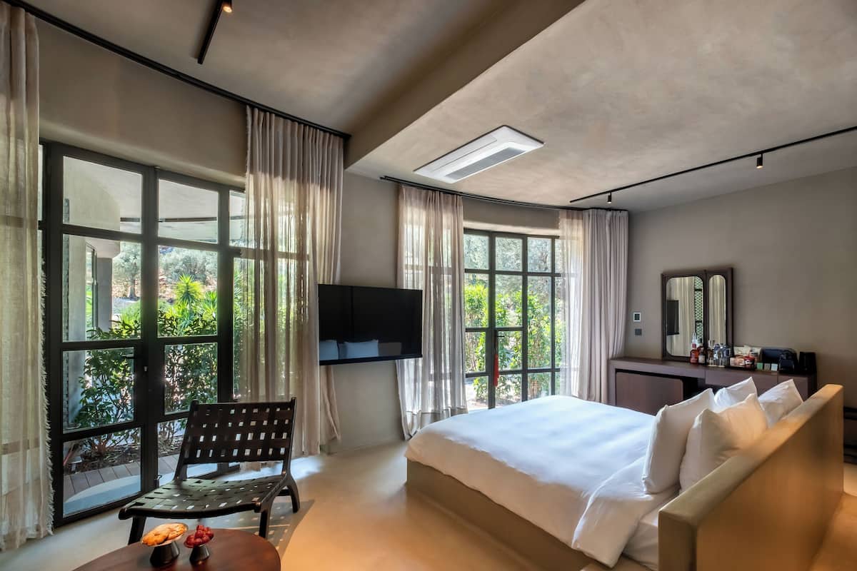 A hotel room with a large bed, a desk with a chair, a TV mounted on the wall, and large windows offering a view of greenery outside.