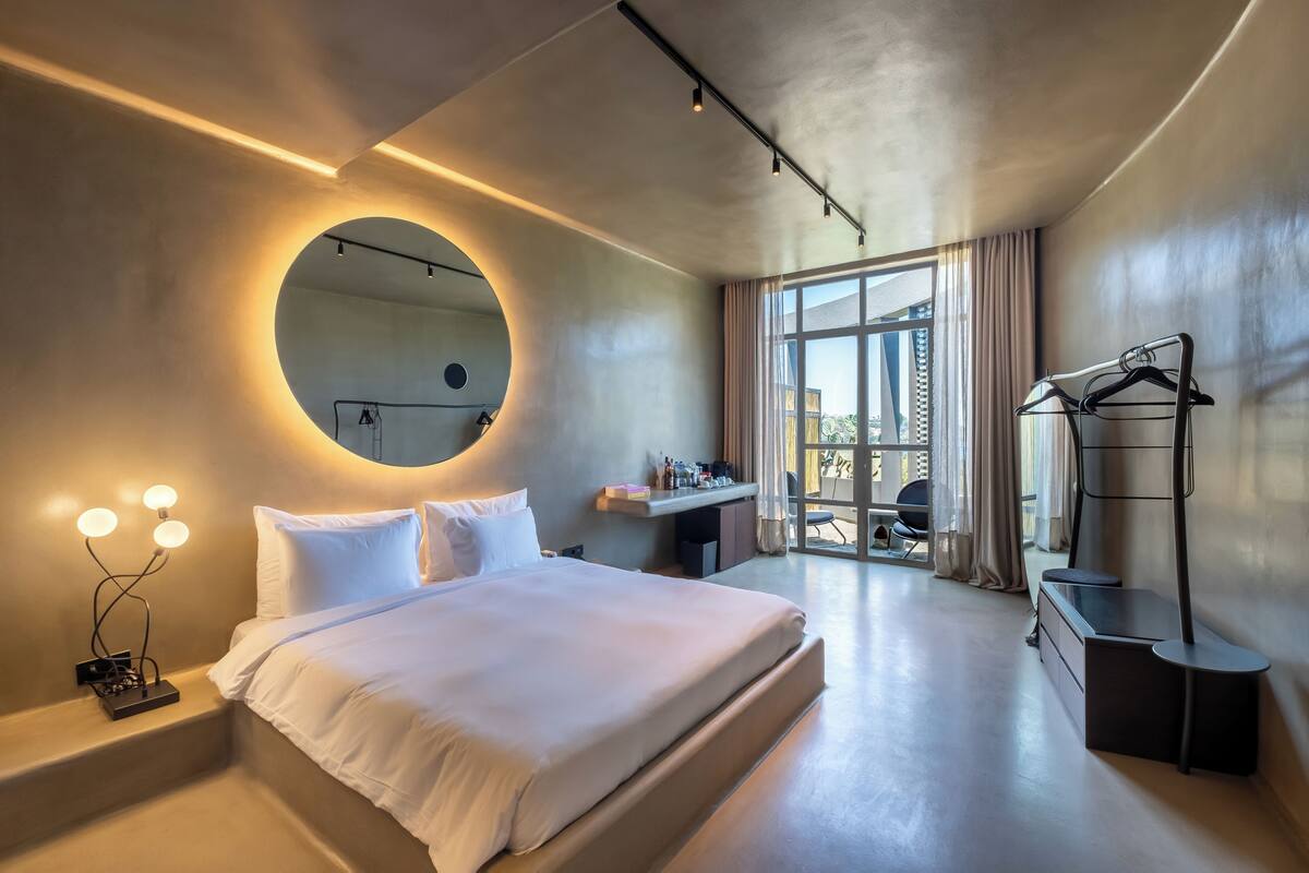 A modern hotel room with a large bed, a round mirror, bedside lighting, a desk, and a wardrobe.