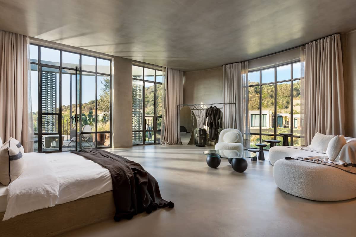 A spacious room with a large bed, a round seating area, and a view of a building through large windows.