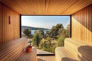 Una sauna de madera con un banco y un balde, con vista a un lago y árboles.
