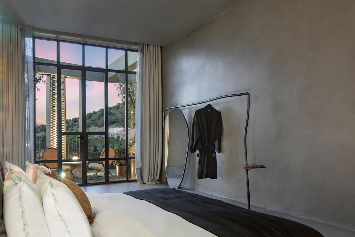 A bedroom with a large bed, a window with curtains, a metal rack with a robe, and a view of a landscape.