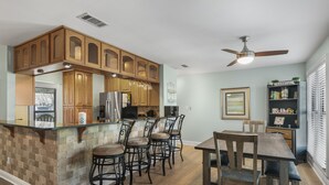 Une cuisine avec des meubles en bois, un coin bar avec des tabourets, une table à manger avec des chaises et un ventilateur de plafond.
