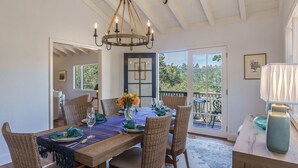 Uma sala de jantar com mesa de madeira posta para seis, cadeiras de vime, um candelabro e vista para um pátio externo através de portas de vidro.