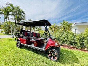 Ein roter Golfwagen steht auf einer Rasenfläche, im Hintergrund sind Palmen und ein weißes Gebäude zu sehen.