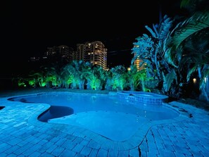 A nighttime view of a swimming pool area with a brick pathway, palm trees, and illuminated buildings in the background.