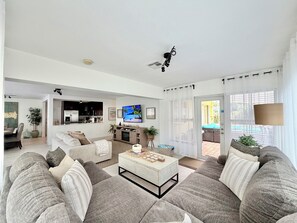 A modern living room with a sofa, coffee table, and a television. There is a kitchen area in the background with cabinets and a dining table.