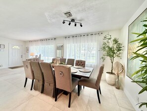 A dining area with a table set for six, surrounded by six chairs, a ceiling light fixture, and a large window with curtains.