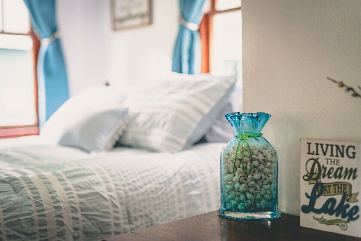 A bed with a white and green patterned bedspread, a blue vase with white pebbles, and a sign that reads "LIVING THE Dream AT THE Lake".