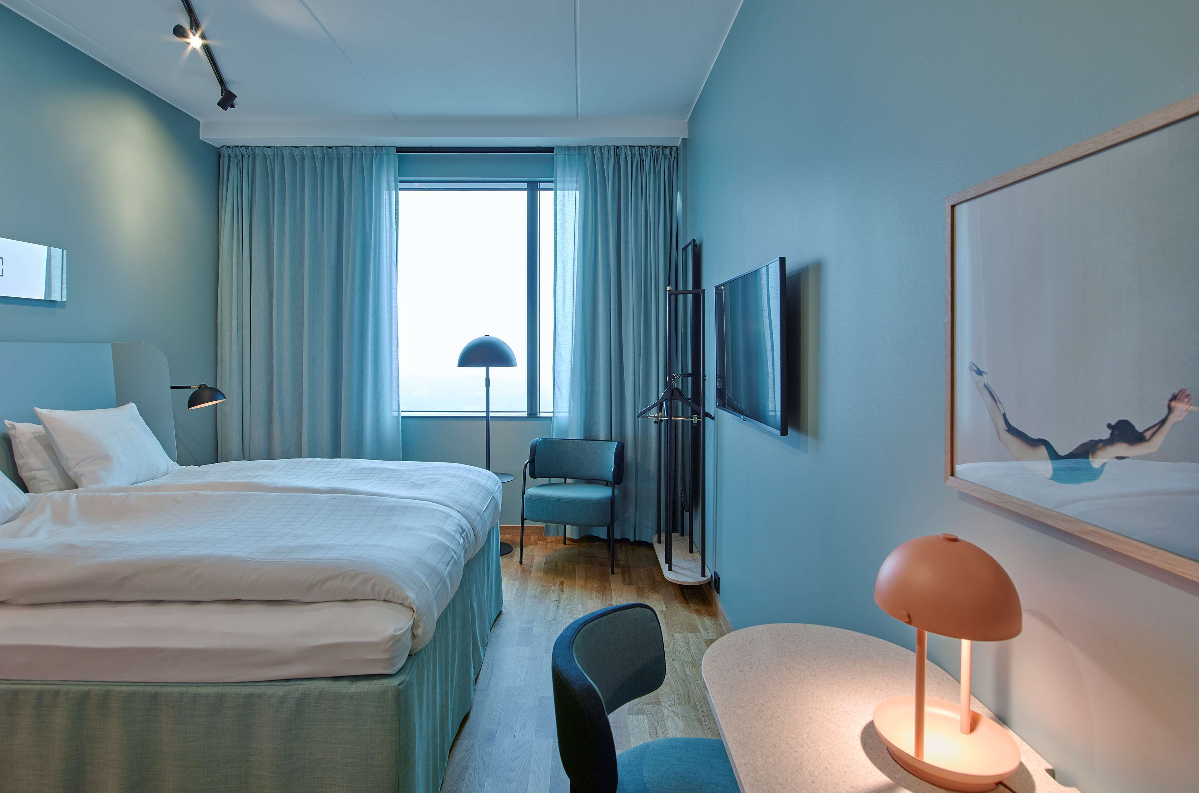 A hotel room with a bed, a desk, a chair, a lamp, a TV, and a window with curtains.