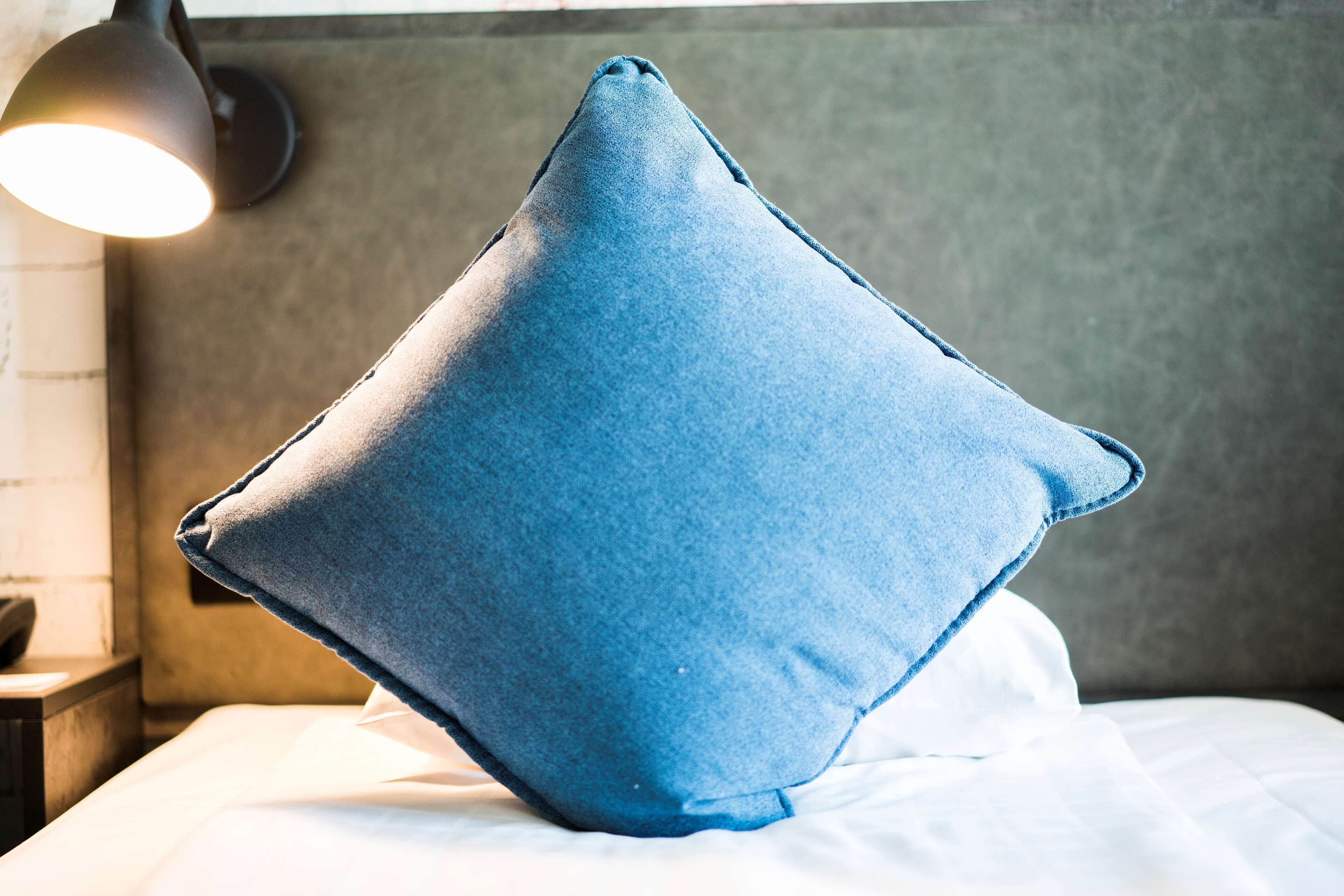 A blue pillow on a bed with a textured wall and a lamp in the background.