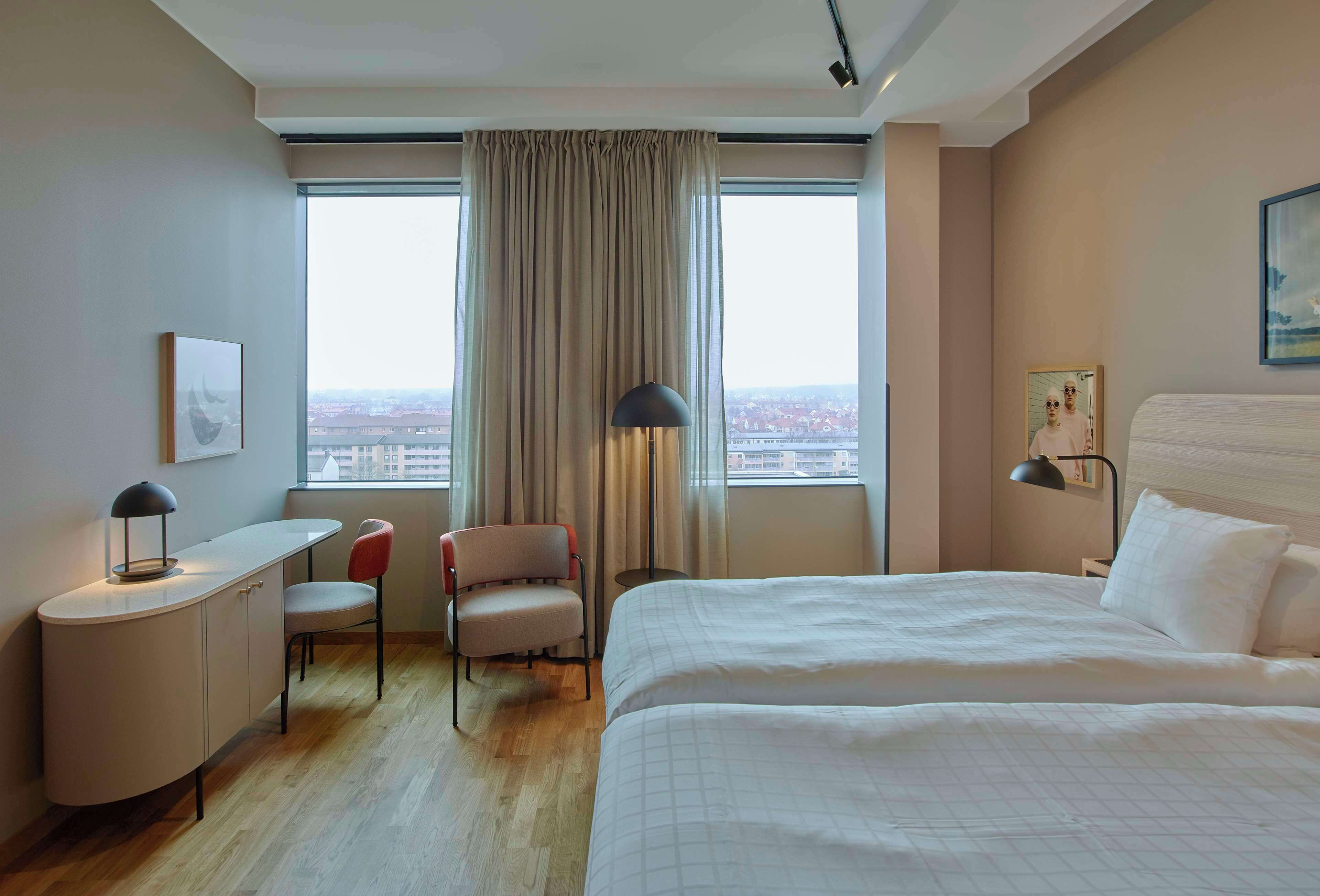Une chambre d’hôtel équipée d’un lit, d’un bureau, d’une chaise et offrant une vue sur la ville grâce à de grandes fenêtres.