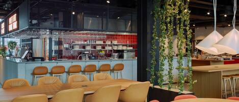 A modern restaurant interior with a long wooden table and several chairs, a bar area with glasses and kitchen equipment, and a vertical garden.