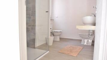 Ein Badezimmer mit Duschkabine aus Glas, zwei Toiletten, einem Waschbecken und einer gewobenen Matte auf dem Boden.