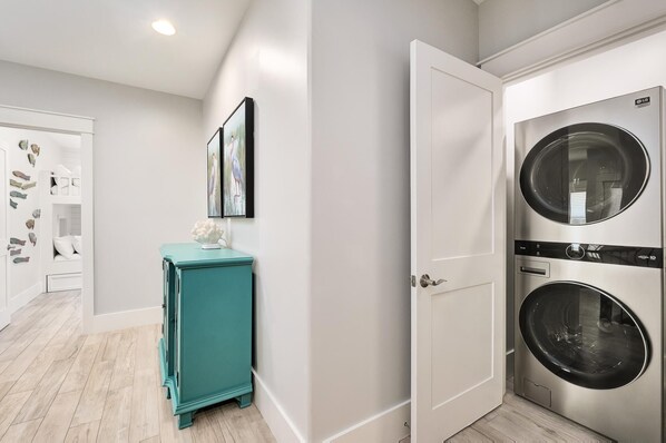 A laundry room with a front-loading washer and dryer, a teal cabinet, and a door leading to a bedroom.