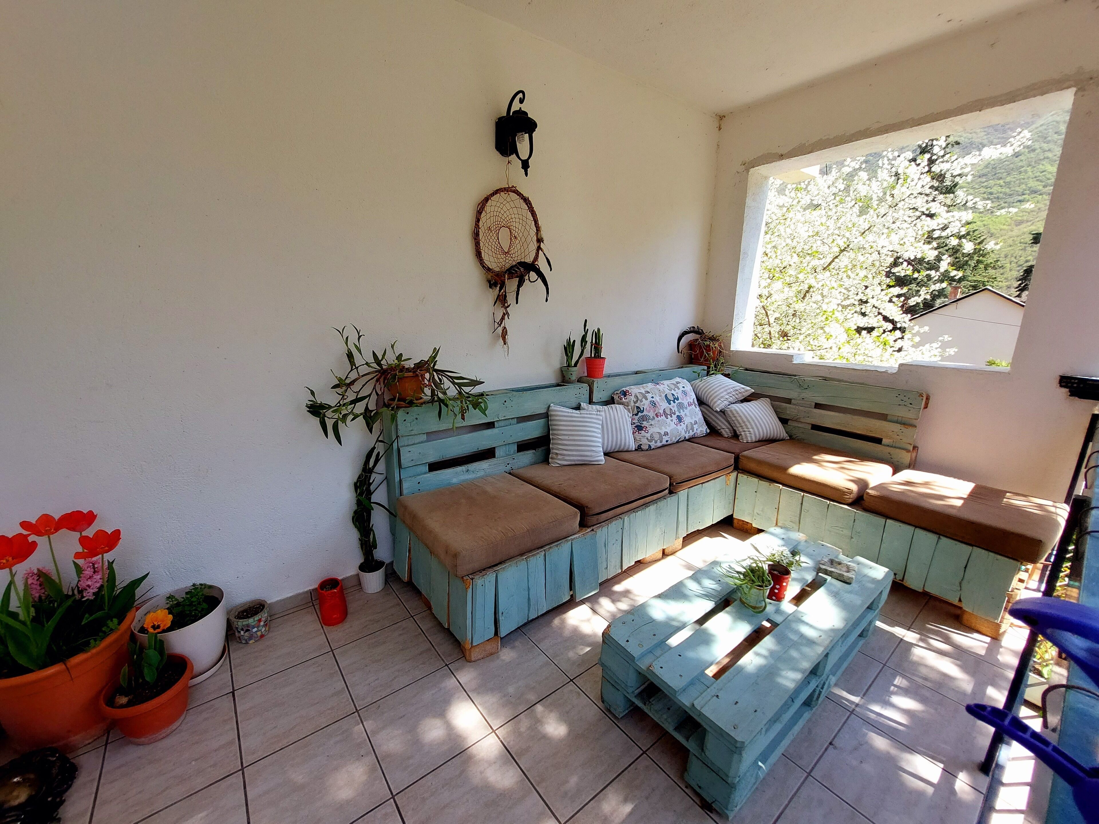 A cozy seating area with a wooden bench and cushions, surrounded by potted plants and a wall-mounted dreamcatcher.
