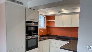 A compact kitchen with white cabinets, a black oven, and a window with orange walls. - LE CLOS DES 2 CAPS - RENOVATED CONTEMPORARY 2 BED APARTMENT 2 STEPS FROM THE SEA (Wimereux)