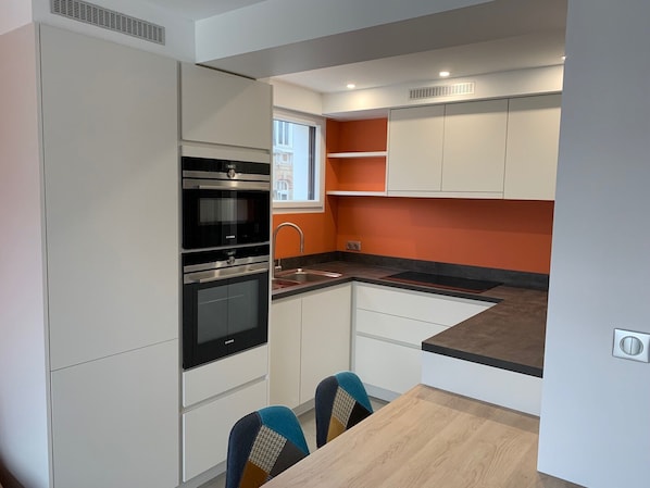 A compact kitchen with white cabinets, a black oven, and a window with orange walls. - LE CLOS DES 2 CAPS - RENOVATED CONTEMPORARY 2 BED APARTMENT 2 STEPS FROM THE SEA (Wimereux)