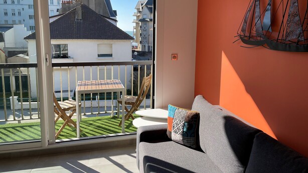 A balcony with a table and chairs, a grey sofa, and a view of buildings and a green lawn. - LE CLOS DES 2 CAPS - RENOVATED CONTEMPORARY 2 BED APARTMENT 2 STEPS FROM THE SEA (Wimereux)
