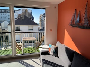 A balcony with a table and chairs, a grey sofa, and a view of buildings and a green lawn. - LE CLOS DES 2 CAPS - RENOVATED CONTEMPORARY 2 BED APARTMENT 2 STEPS FROM THE SEA (Wimereux)