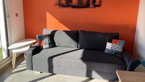 A modern living room with a grey sofa, two patterned throw pillows, a small white side table, and a wall-mounted abstract art piece. - LE CLOS DES 2 CAPS - RENOVATED CONTEMPORARY 2 BED APARTMENT 2 STEPS FROM THE SEA (Wimereux)