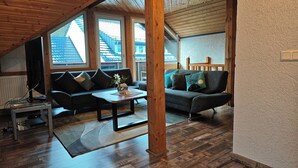 A loft-style living room with wooden beams, two sofas, a coffee table, and large windows offering views of neighboring buildings. - Apartments Zur alten Post (Willingen)
