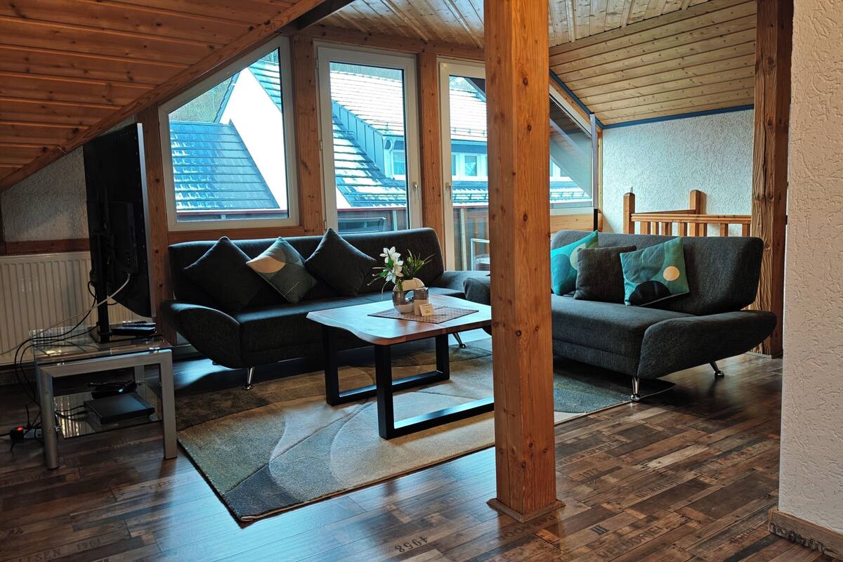 A loft-style living room with wooden beams, two sofas, a coffee table, and large windows offering views of neighboring buildings.