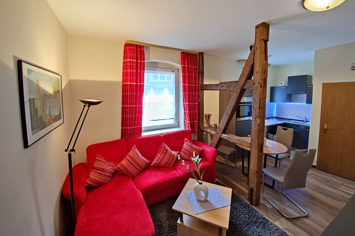 A living room with a red sofa, a wooden ladder, a dining table with chairs, and a kitchen area visible through an open door.