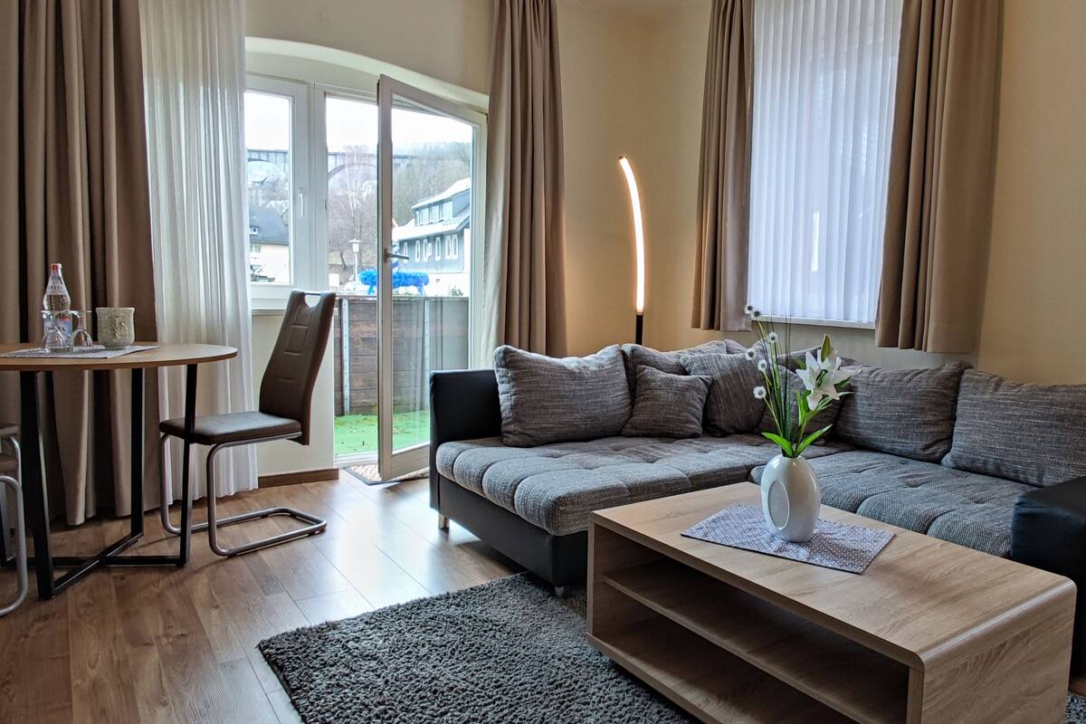 A modern living room with a grey sofa, a wooden coffee table, and a dining area with a table and chairs.