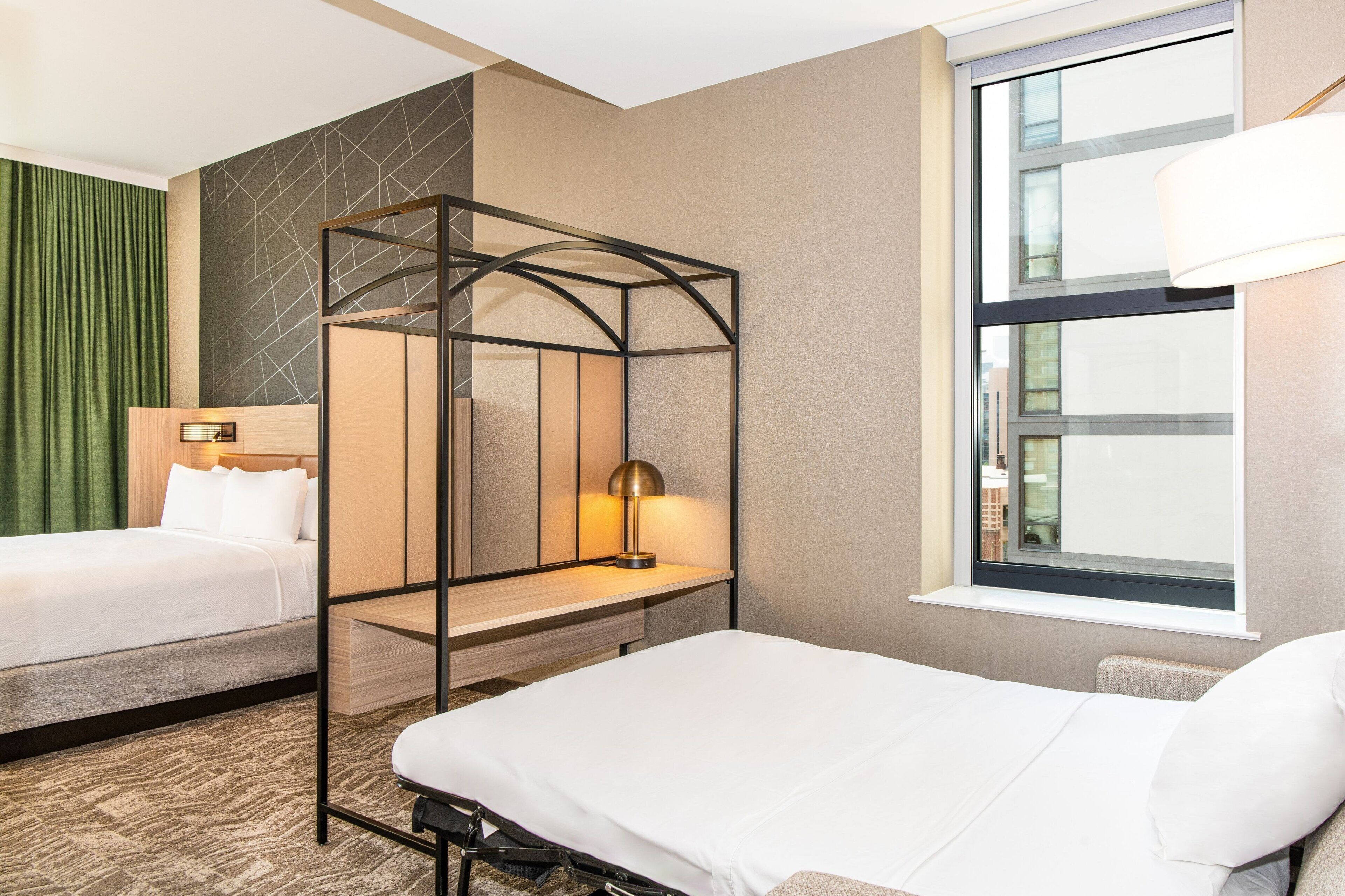 A hotel room with two beds, a standing desk, a window with a view, and a lamp.