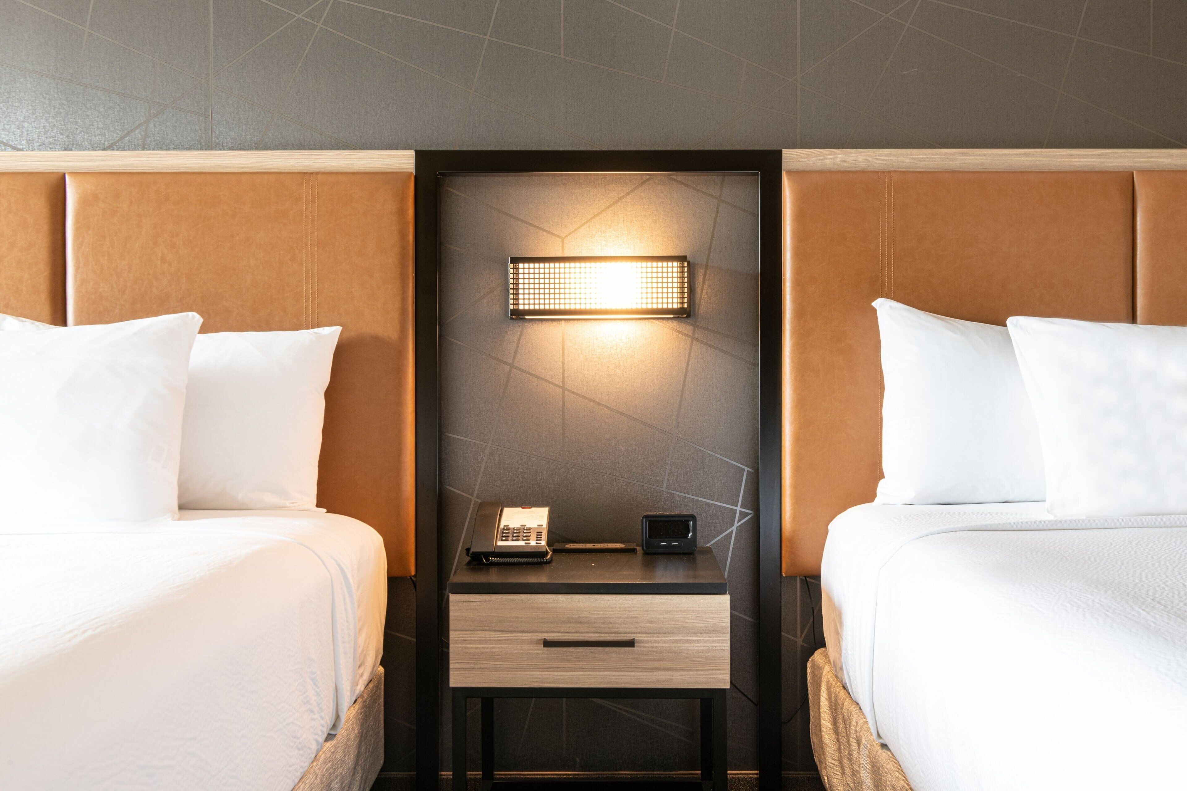 A hotel room with two beds, a nightstand with a lamp, and a wall-mounted light fixture.