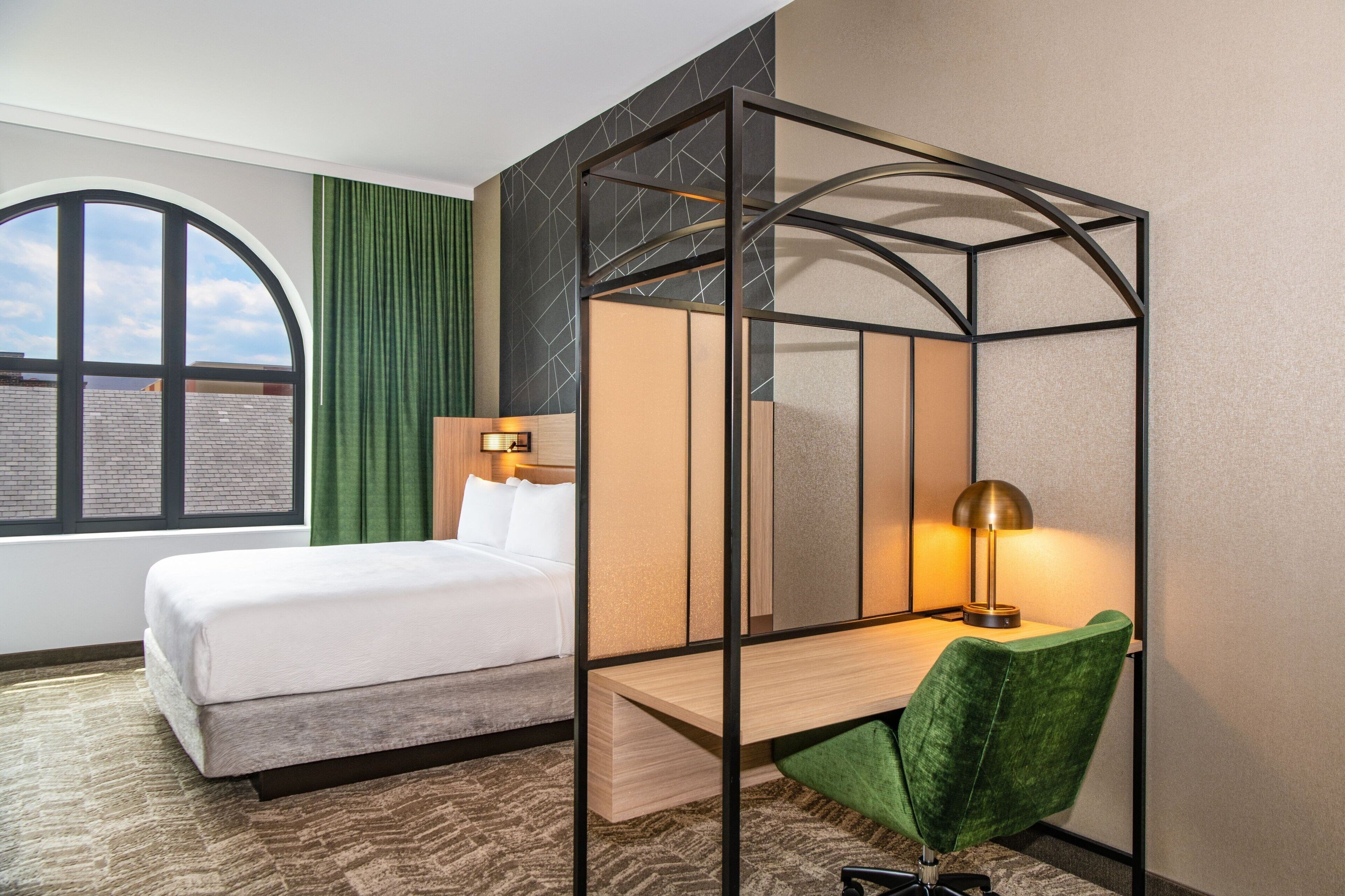 Una habitación de hotel con una cama, un escritorio con una silla, una ventana con cortinas y vistas a la ciudad.