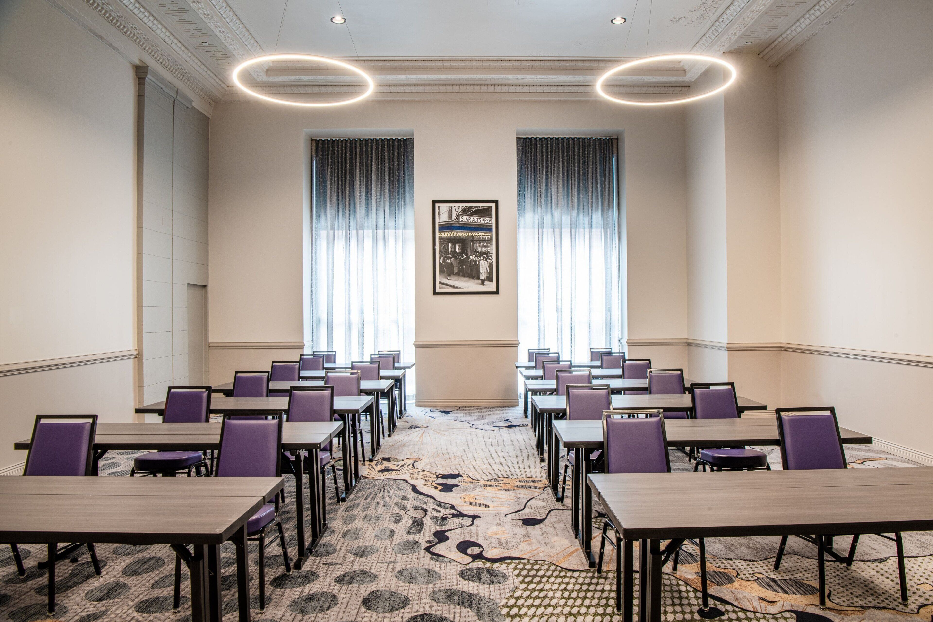 A conference room with rows of tables and chairs, large windows with curtains, and a patterned carpet.