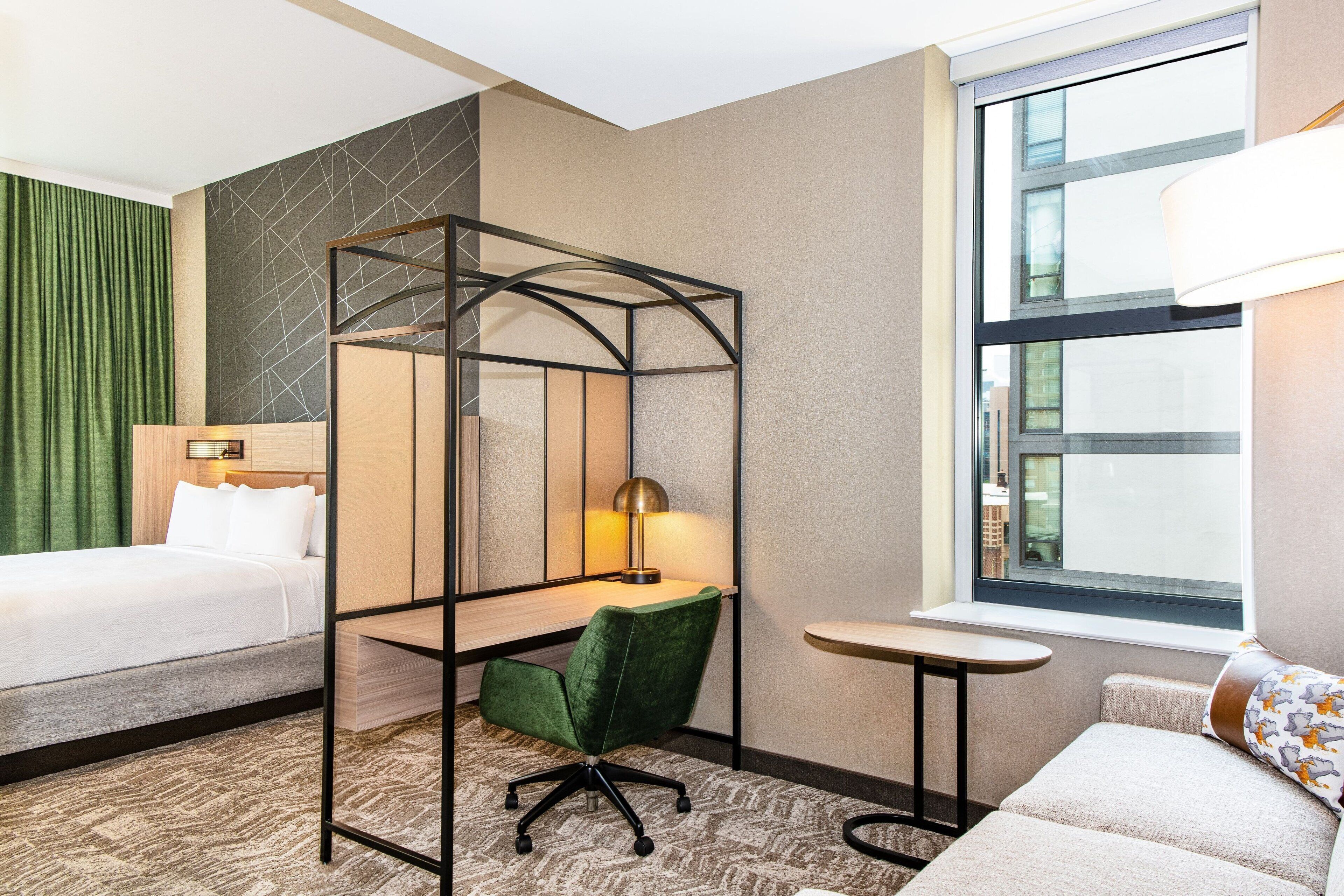 Habitación de hotel con cama, escritorio con silla, una mesita, un sofá y ventana con vistas a edificios.
