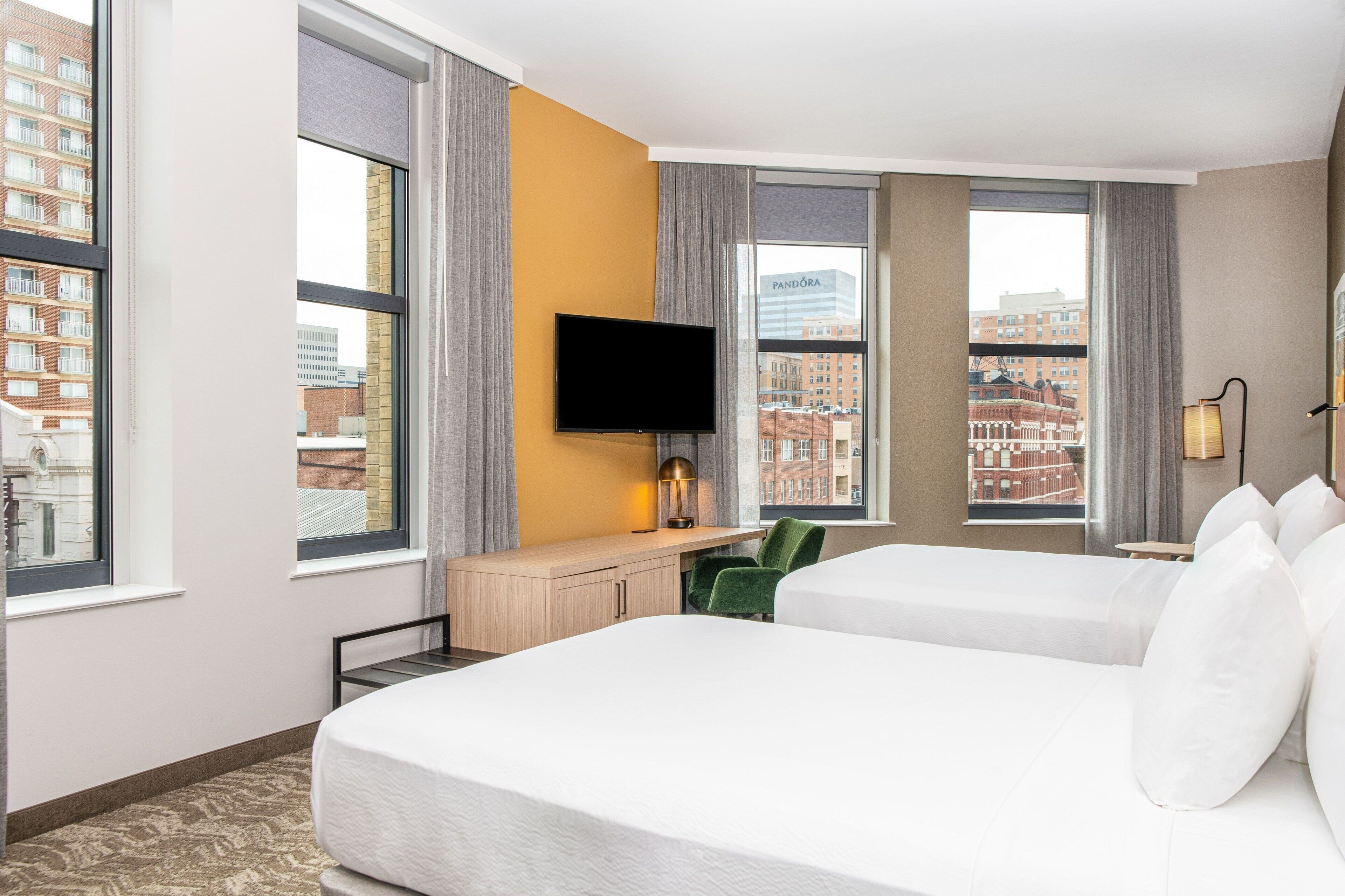 A hotel room with two beds, a desk with a flat-screen TV, a chair, and large windows offering city views.