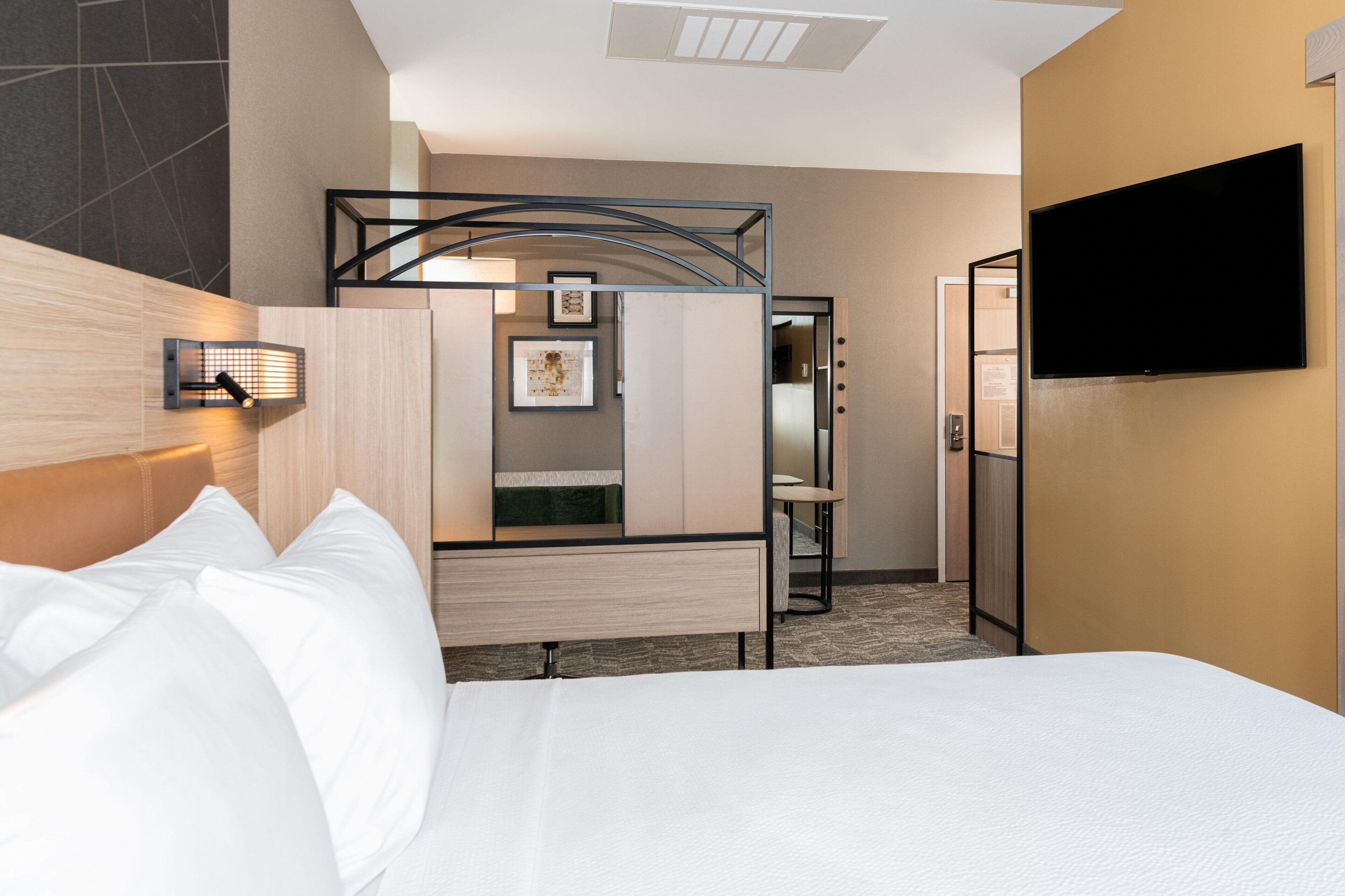 A hotel room with a bed, a TV mounted on the wall, a desk with a chair, and a mirror.