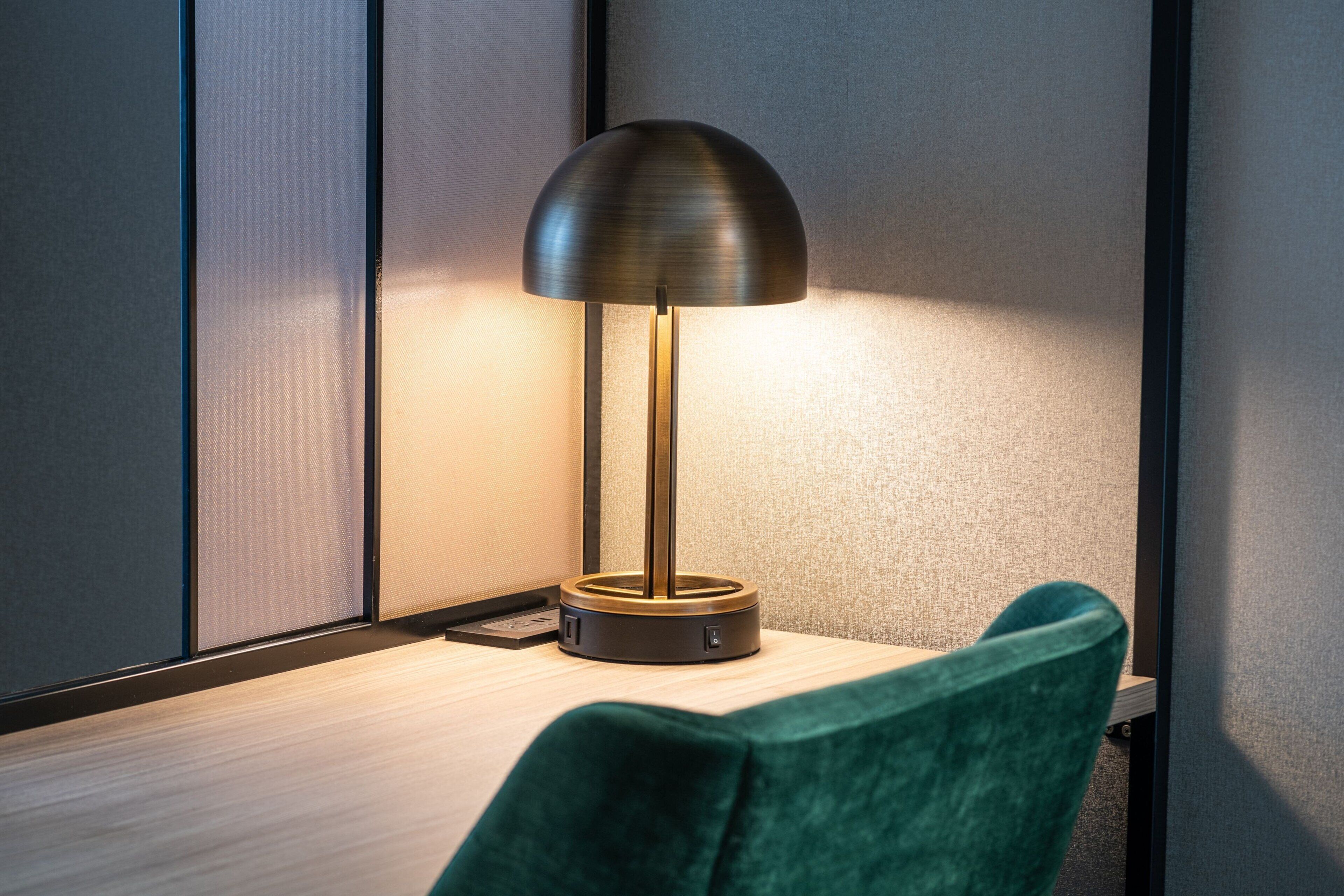 Uma luminária de mesa moderna com base metálica e cúpula curva, sobre uma escrivaninha de madeira em frente a uma parede texturizada.