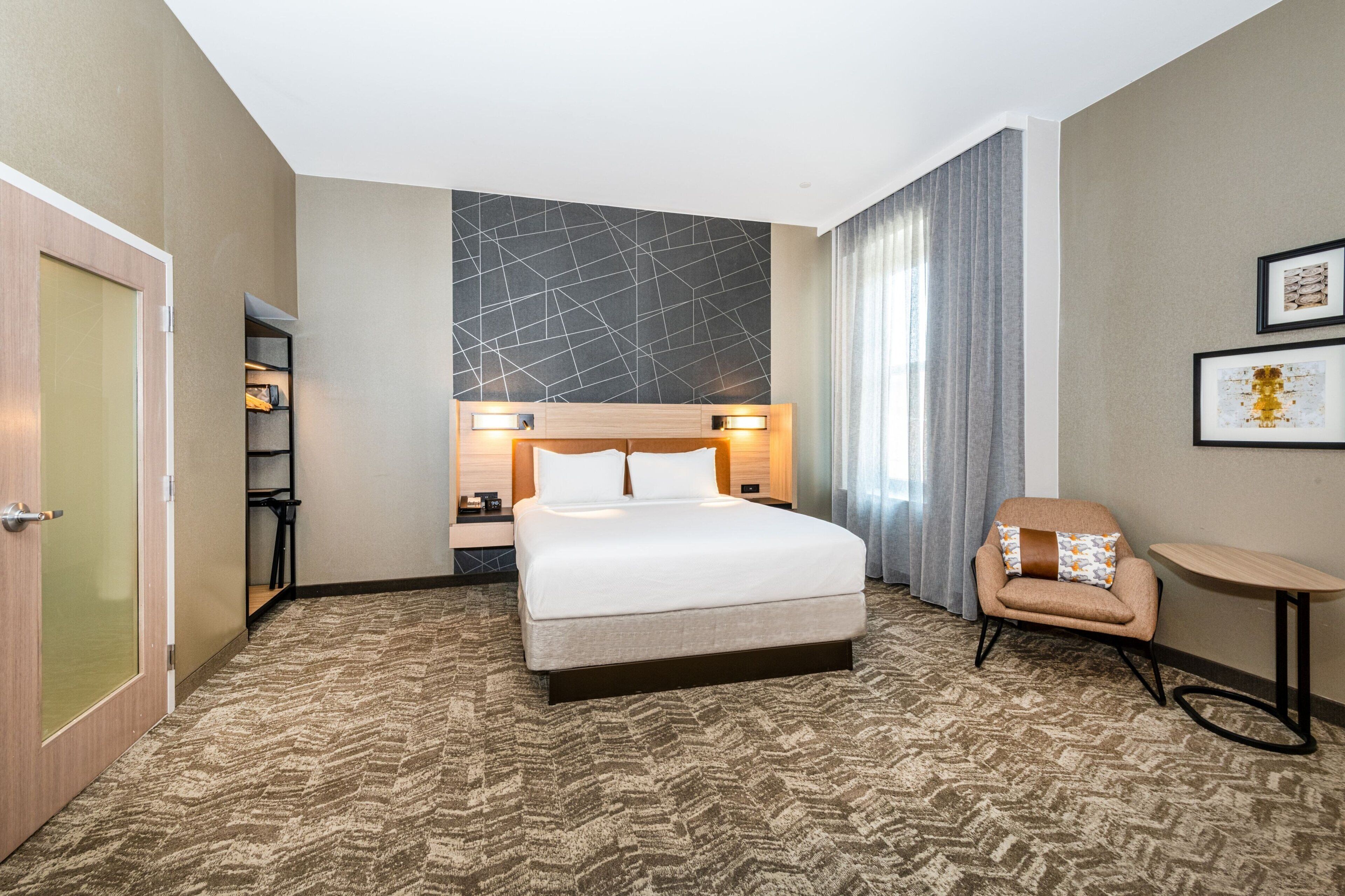 A hotel room with a large bed, a chair, a small table, and a wall-mounted shelf.