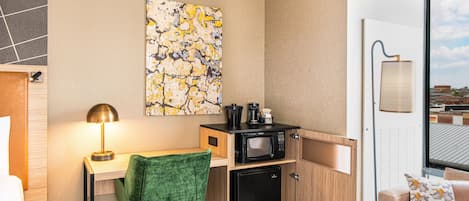 Ein kompaktes Hotelzimmer mit Küchenzeile, einem grünen Sessel, einem kleinen Kühlschrank, einer Mikrowelle, einer Kaffeemaschine und einem großen Fenster mit Blick auf die Stadt.
