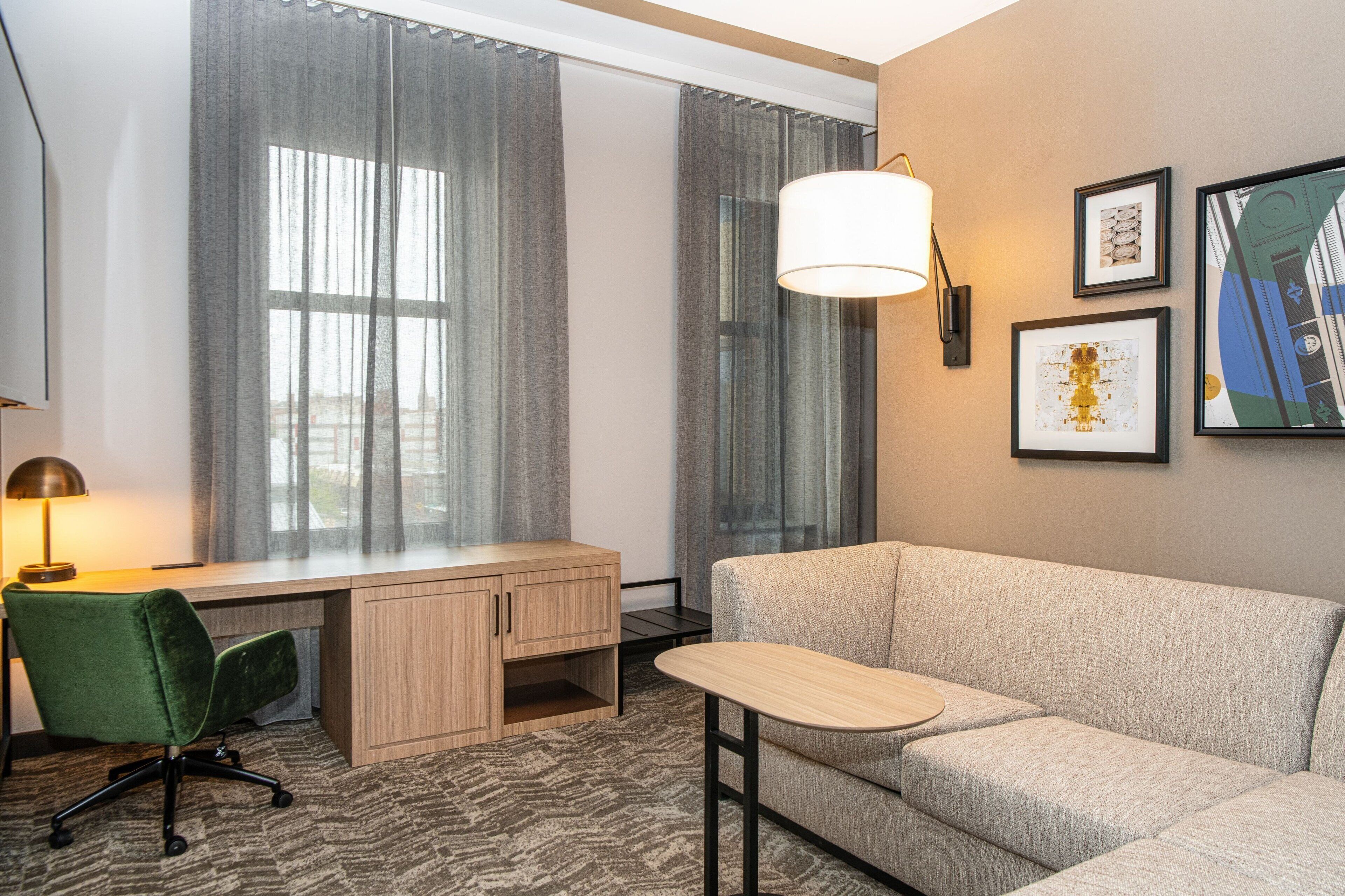 Una habitación de hotel moderna con escritorio, una silla verde, sofá y una obra de arte en la pared.