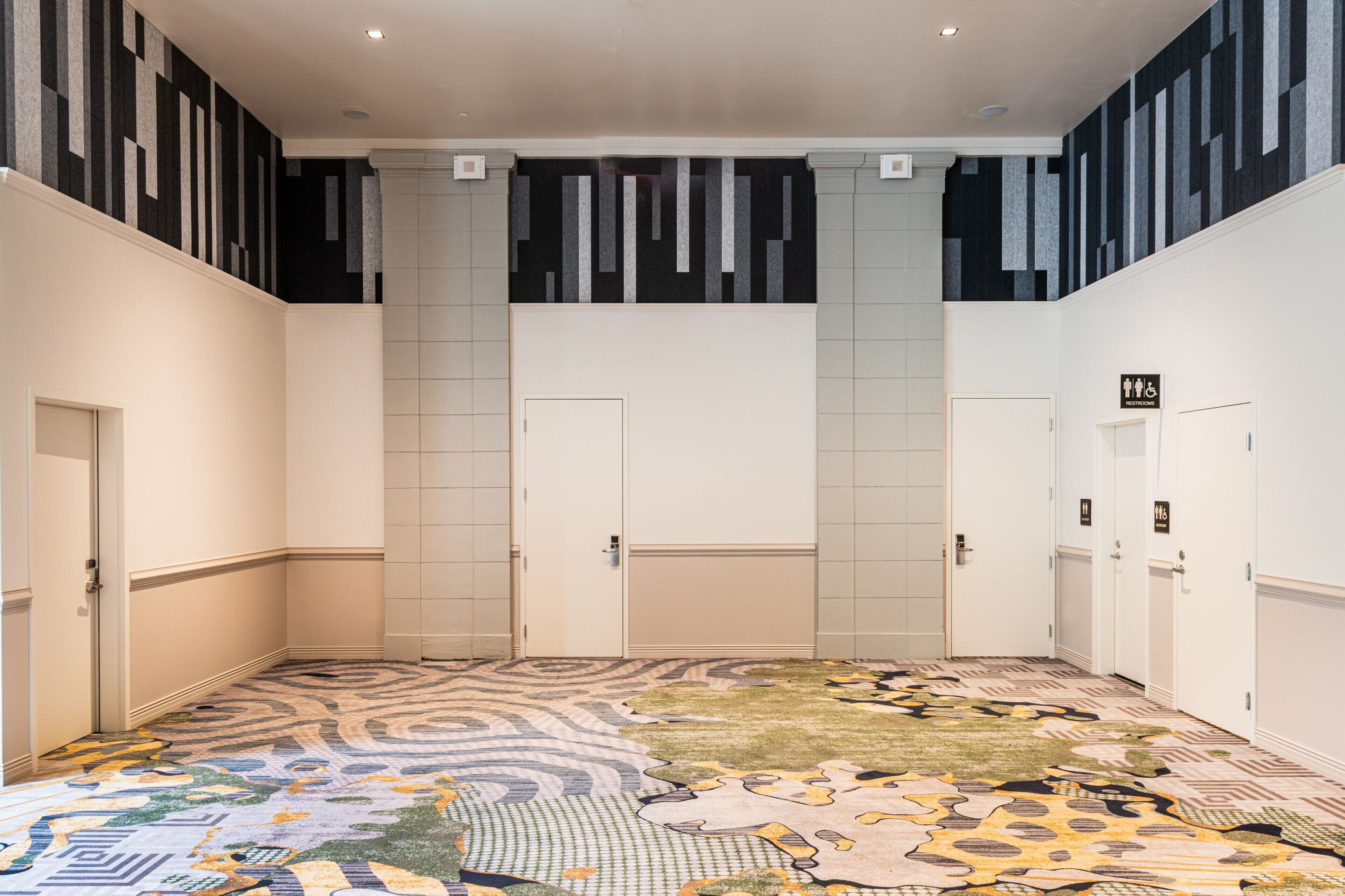 Un hall d’hôtel avec un tapis à motifs, des murs blancs et des portes portant une indication signalant les toilettes pour hommes.