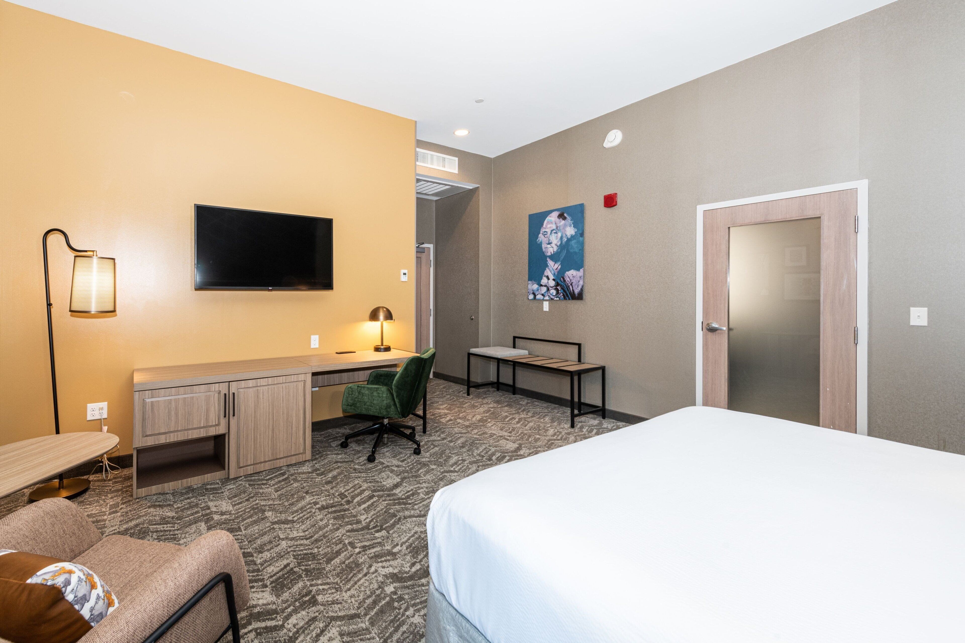A hotel room with a bed, desk, chair, TV, and artwork on the wall.