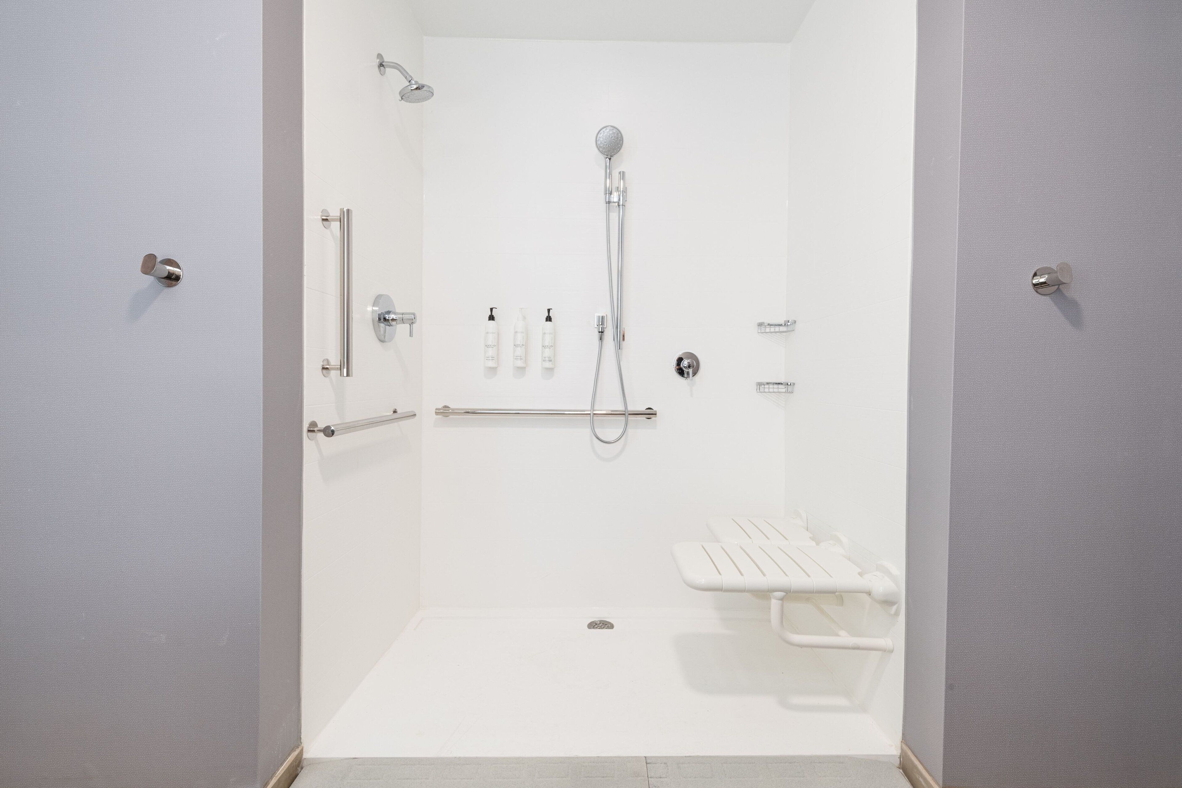 Une salle de douche blanche et accessible, équipée d’un pommeau de douche à main, de barres d’appui et d’un banc.