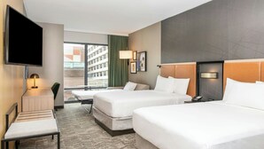 Ein Hotelzimmer mit zwei Betten, einem Schreibtisch, einem Fernseher und einem Fenster mit Blick auf Gebäude.