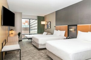 Habitación de hotel con dos camas, un escritorio, un televisor y una ventana con vistas a edificios.