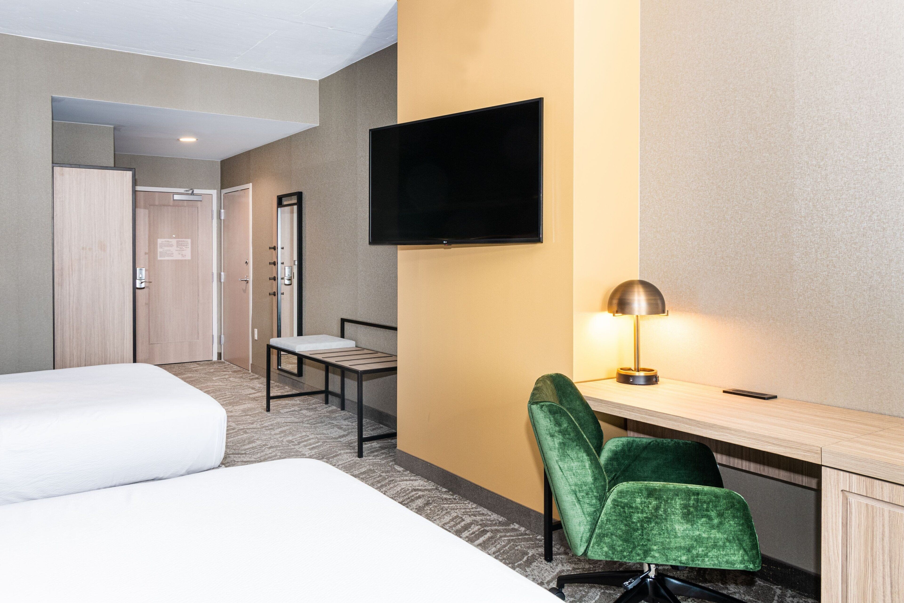 Ein Hotelzimmer mit zwei Betten, einem Schreibtisch mit Lampe, einem an der Wand befestigten Fernseher und einem Sessel.