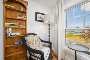 A room with a wicker chair, a wooden bookshelf, a framed picture, a floor lamp, and a view of a body of water through a window. - Island Sunset by Seaside Vacations & Sales (Chincoteague Island)