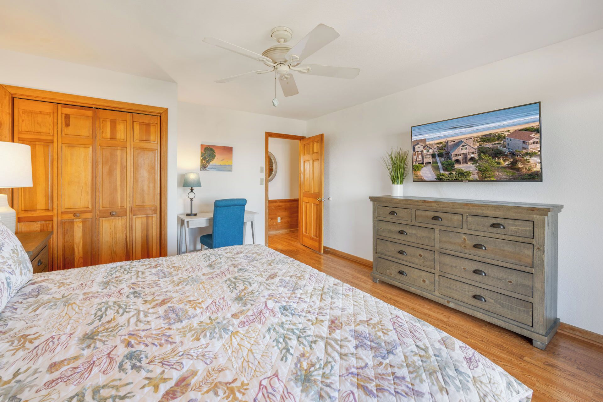 A bedroom with a bed, a desk, a chair, a dresser, a ceiling fan, and a painting of a coastal town.