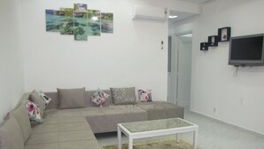 Una sala de estar con un sofá color beige, una mesa de centro y un tapete estampado. Hay tres cuadros en la pared y un televisor montado.
